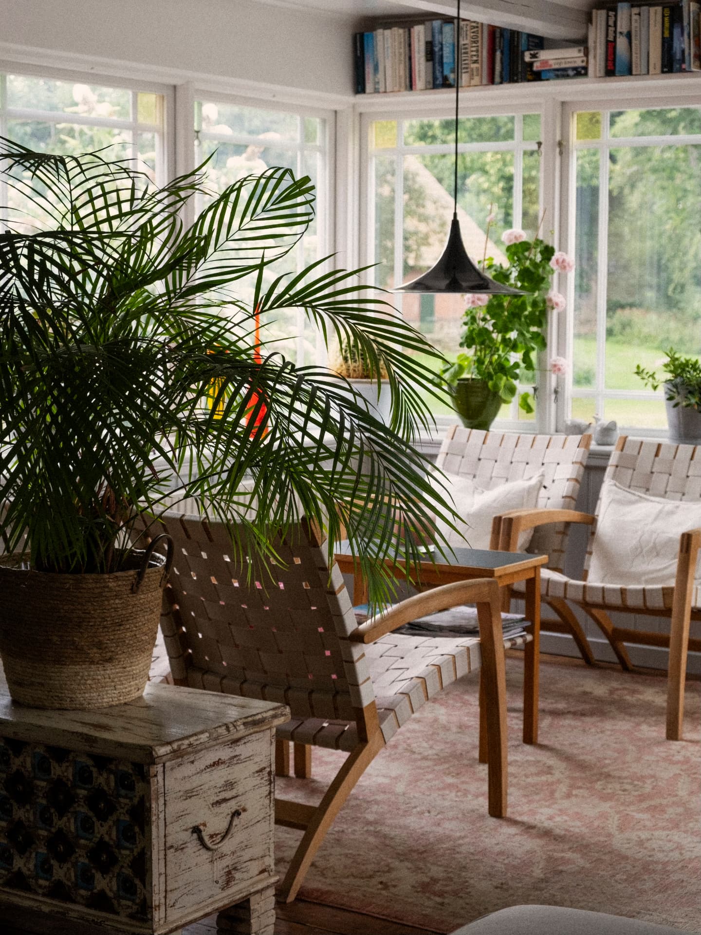 Ljus salong med flätade fåtöljer, palm och stora spröjsade fönster mot trädgården
